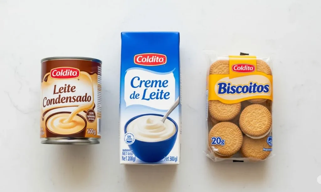 Foto de cima (flat lay) de uma lata de leite condensado, uma embalagem de creme de leite e um pacote de biscoitos alinhados sobre uma superfície clara, representando os três ingredientes principais para uma sobremesa de travessa simples.