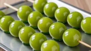 Uma imagem vibrante de Uvas do Amor verdes brilhantes, revestidas de açúcar, em espetos, arranjadas em uma bandeja metálica sob iluminação de estúdio. O foco está no revestimento de açúcar liso e brilhante.